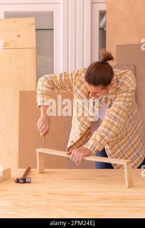 Crop faceless male carpenter screwing bolt with screwdriver into wooden plank on table near hammer while assembling furniture in workshop Stock Photo