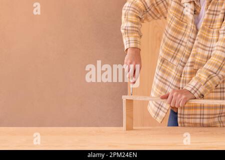 Crop faceless male carpenter screwing bolt with screwdriver into wooden plank on table near hammer while assembling furniture in workshop Stock Photo
