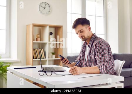 Smiling businessman holding smartphone sitting in front of laptop Stock Photo