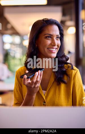 Mature Businesswoman Working On Laptop At Desk In Office Talking Into Mic Of Mobile Phone Stock Photo