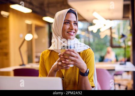 Mature Businesswoman Wearing Headscarf Working On Laptop At Desk In Office With Takeaway Drink Stock Photo