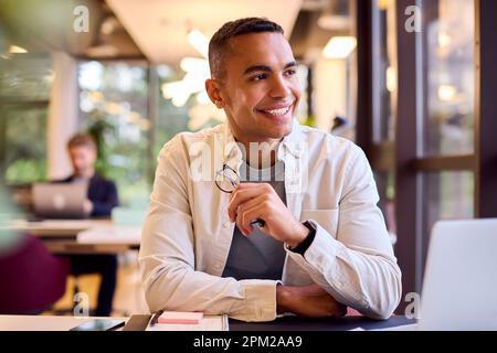 Young Businessman Working On Laptop At Desk In Office Talking Into Mic Of Mobile Phone Stock Photo