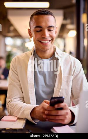 Young Businessman Working On Laptop At Desk In Office Checking Messages On Mobile Phone Stock Photo