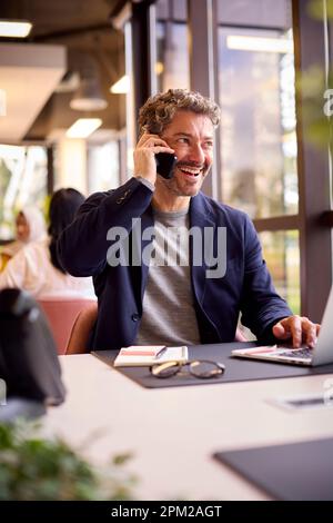 Mature Businessman Working On Laptop At Desk In Office Talking On Mobile Phone Stock Photo