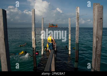 Man on jetty wearing life jacket about to get into water to join other snorkellers with oil rig in background, Sipadan Water Village, Mabul Island, Sa Stock Photo