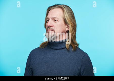 Portrait of mature bearded man with long hair looking away standing against blue background Stock Photo