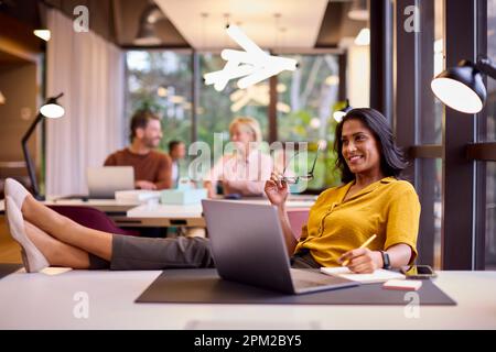 Mature Businesswoman With Feet On Desk Working On Laptop In Office Stock Photo
