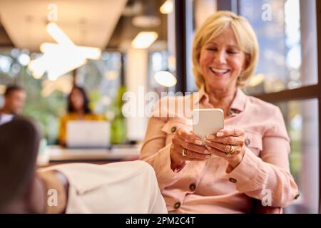 Mature Businesswoman With Feet On Desk Working In Office Checking Messages On Mobile Phone Stock Photo