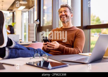 Casually Dressed Mature Businessman With Feet On Desk In Office Using Mobile Phone Stock Photo