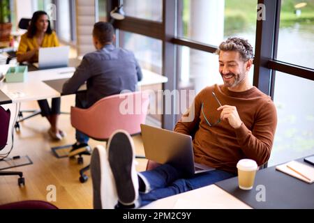 Casually Dressed Mature Businessman With Feet On Desk In Office Working On Laptop Stock Photo
