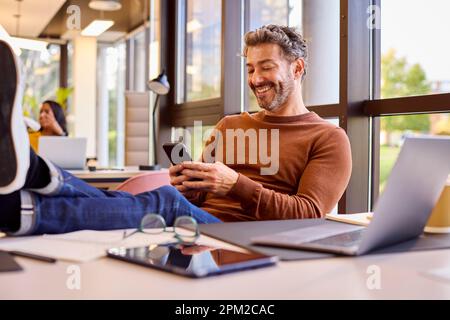 Casually Dressed Mature Businessman With Feet On Desk In Office Using Mobile Phone Stock Photo