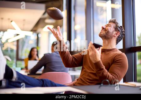 Mature Businessman With Feet On Desk In Office Talking Into Mic Of Mobile Phone Catching Football Stock Photo