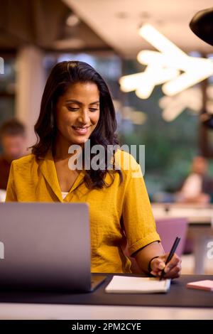 Mature Businesswoman Working On Laptop At Desk In Office Writing In Notebook Stock Photo