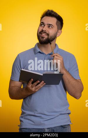 Thoughtful journalist caucasian man making notes, writing down thoughts with pen into notepad notebook diary, to do list, good idea. Handsome bearded guy isolated alone on yellow studio background Stock Photo
