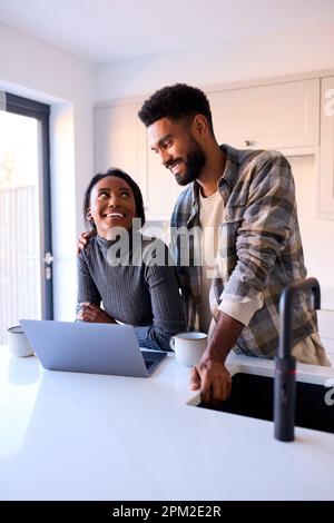 Young Couple At Home Looking At Laptop On Counter In Kitchen Together Stock Photo