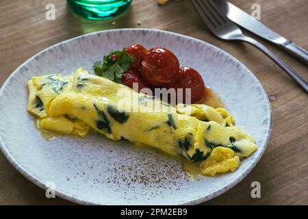 Homemade omelet with spinach and roasted cherry tomatoes in ceramic plate on rustic wooden table, healthy breakfast French omelette with greens Stock Photo