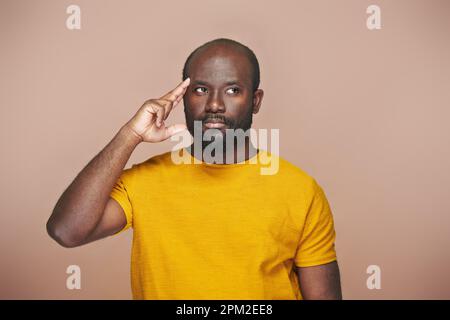 Portrait of African American pensive man in yellow shirt pointing at his head standing on brown background Stock Photo