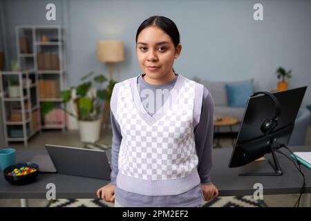 Waist up portrait of Latin American young woman looking at camera while standing by home office workplace Stock Photo
