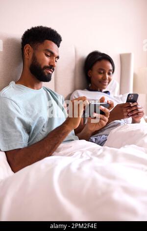 Young Couple At Home Both Looking At Mobile Phones In Bed Together Stock Photo