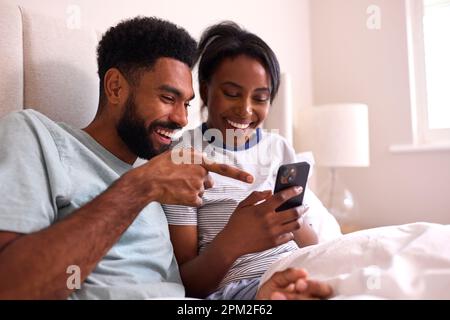 Young Couple At Home Both Looking At Mobile Phones In Bed Together Stock Photo