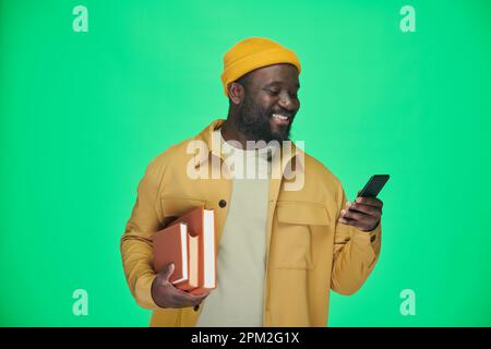 African American man with books reading message on his mobile phone and smiling while standing on green background Stock Photo