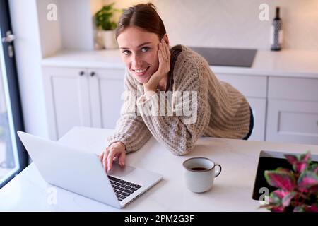 Portrait Of Young Woman At Home Working On Laptop On Counter In Kitchen Stock Photo