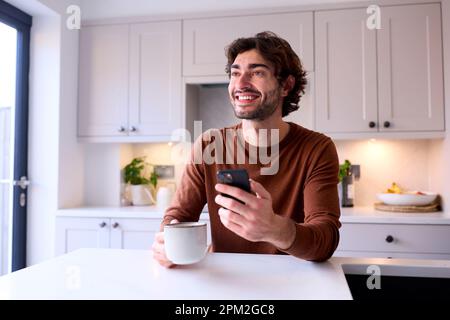 Young Man Relaxing At Home In Kitchen Holding Hot Drink Using Mobile Phone Stock Photo