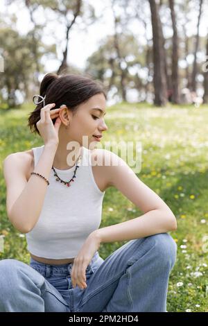 Side view of young woman putting daisy flower in hair while sitting on lawn in park,stock image Stock Photo