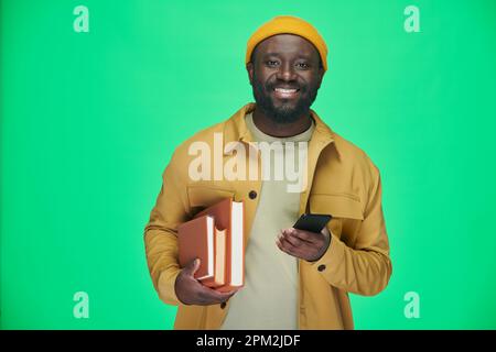 Portrait of African American man with books smiling at camera while using his smartphone against green background Stock Photo
