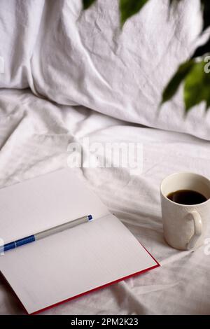 An open notebook with a cup of coffee on a bed with white sheets Stock Photo