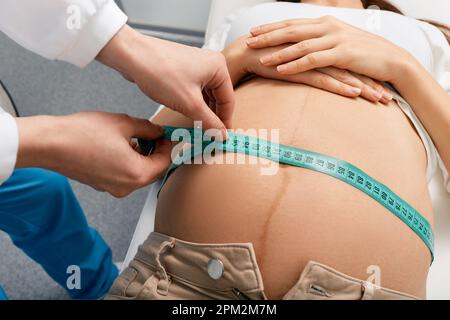 Obstetrician gynecologist with centimeter tape measuring belly of pregnant woman lying on medical bed in hospital, close-up Stock Photo