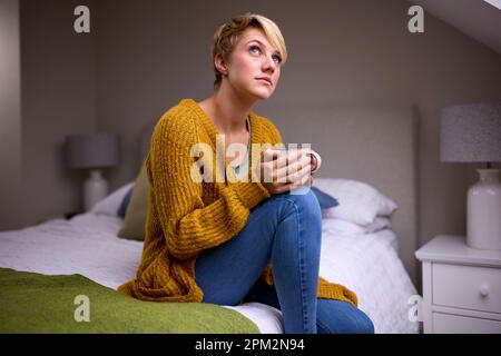 Thoughtful Young Woman With Hot Drink Sitting On Bed At Home Stock Photo