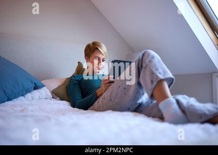 Young Woman Lying On Bed Wearing Pyjamas At Home Looking At Digital Tablet Stock Photo