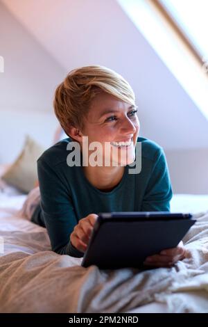 Young Smiling Woman Lying On Bed Wearing Pyjamas At Home Looking At Digital Tablet Stock Photo