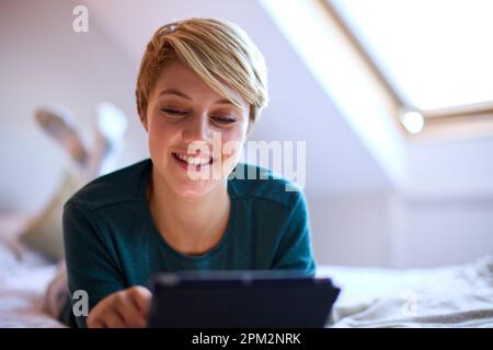 Young Smiling Woman Lying On Bed Wearing Pyjamas At Home Looking At Digital Tablet Stock Photo