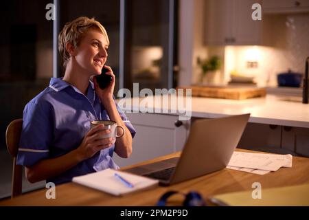 Woman In Medical Scrubs Talking On Mobile Phone Working Or Studying On Laptop At Home At Night Stock Photo