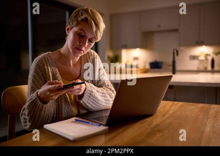 Young Woman Working Or Studying On Laptop At Home At Night Taking Picture Of Notes With Mobile Phone Stock Photo