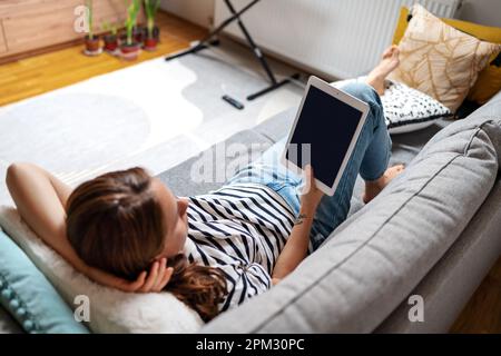 Woman lying down on sofa at home and reeding ebook. Stock Photo