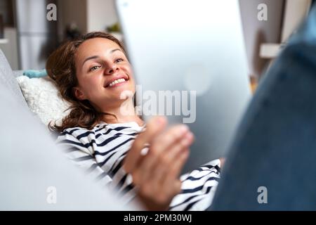 Woman smiling and using her digital tablet at home lying down on sofa. Stock Photo