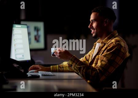 Businessman Working Late In Office With Face Illuminated By Computer Screen Drinking Coffee Stock Photo