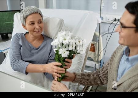 Senior sick woman getting flowers from her son while he visiting her in hospital ward Stock Photo