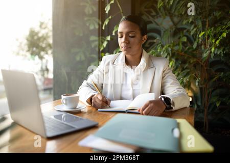 Serious millennial african american businesswoman in white suit working on laptop making notes, planning day Stock Photo