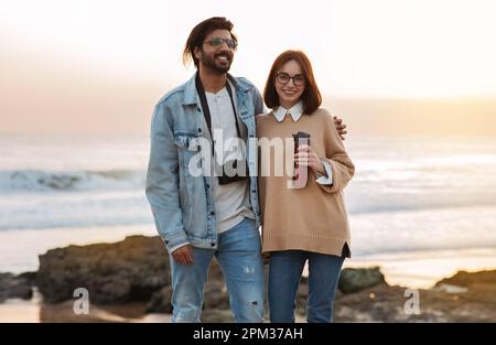 Glad millennial indian male hugging caucasian lady with thermos enjoy ocean, walking on beach outdoor Stock Photo