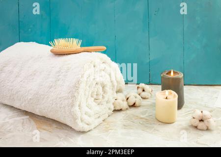 Rolled white towel, wooden hair brush, cotton flowers and lit candles on blue wooden boards and marble background Stock Photo