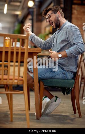 Handsome young man siting in cafe, playing music and dancing to it, enjoying good music. Lifestyle, motivation, lesiure concept. Stock Photo