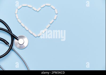 Flat lay. A heart shape made from white medical pills near a doctor's stethoscope on isolated blue background. Ad space Stock Photo