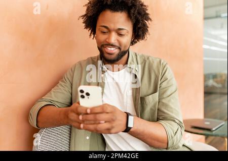 Portrait of smiling man sitting on chair in office, using mobile phone for checking social nets, typing message for family, girlfriend, business partner, looking photos, watches videos Stock Photo