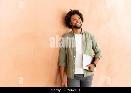 Charismatic smiling positive businessman stands with laptop computer over beige wall, confident small business owner looking distance, male office employee smiling Stock Photo