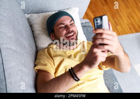Cheerful man messaging online using his smartphone while relaxing on sofa at home. Stock Photo