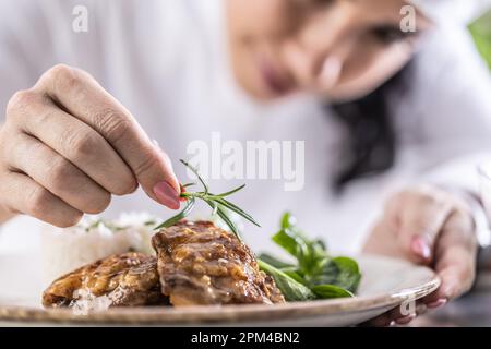 Close-up view of chef's fingers with rosemary sprig decorating food in restaurant kitchen. Stock Photo